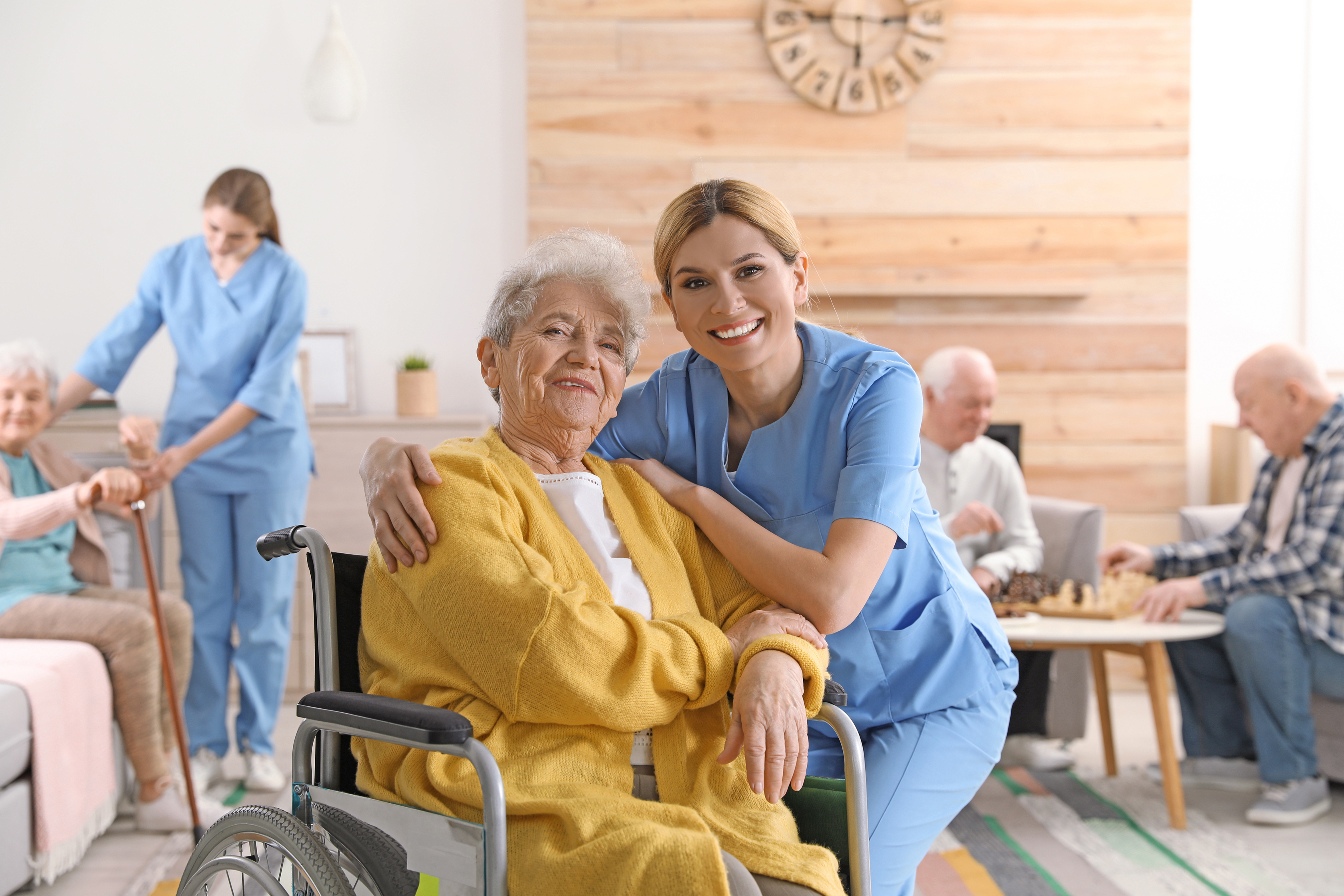 A smiling caregiver poses with an elderly woman in a wheelchair. In the background, other senior individuals are playing chess and walking with assistance, in a cosy communal living space.